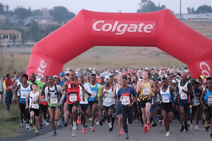 A large group of athletes run from the Colgate Road Race starting line marked with a large Colgate arch